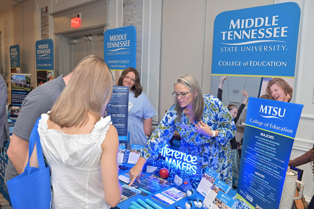 A woman standing behind a table points out some information on a flyer to students standing in front of her.