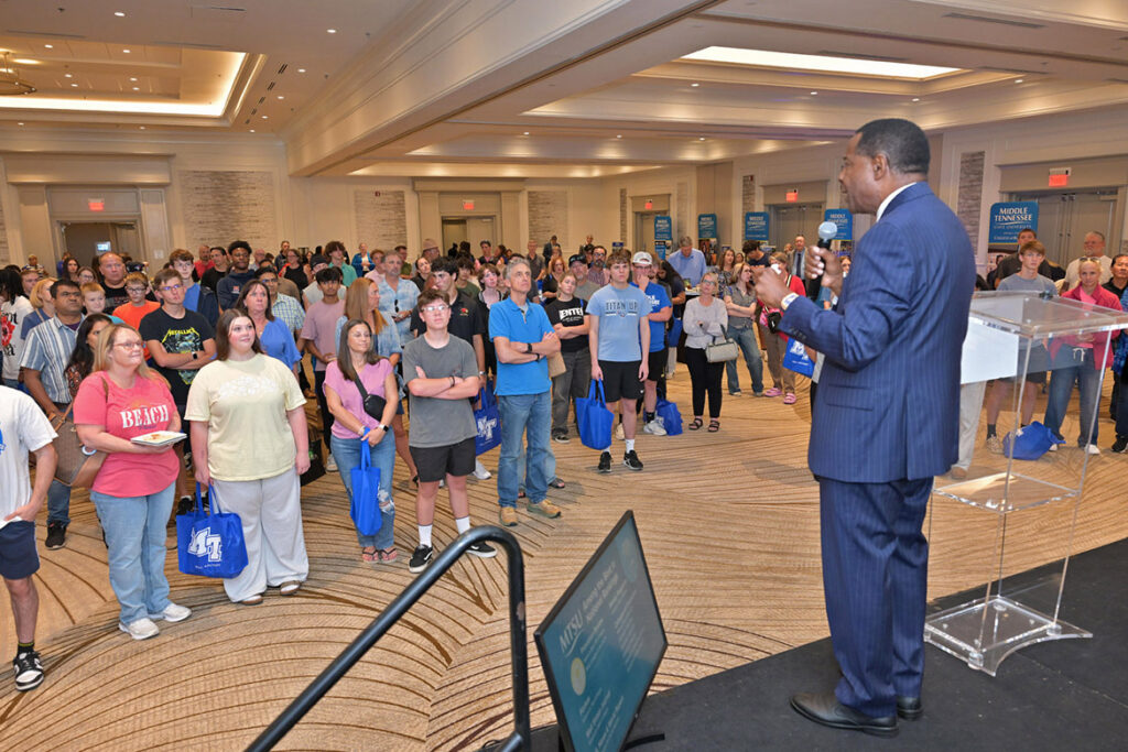 A man in a dark blue, pinstriped suit stands on a stage in front of an audience of students and parents.