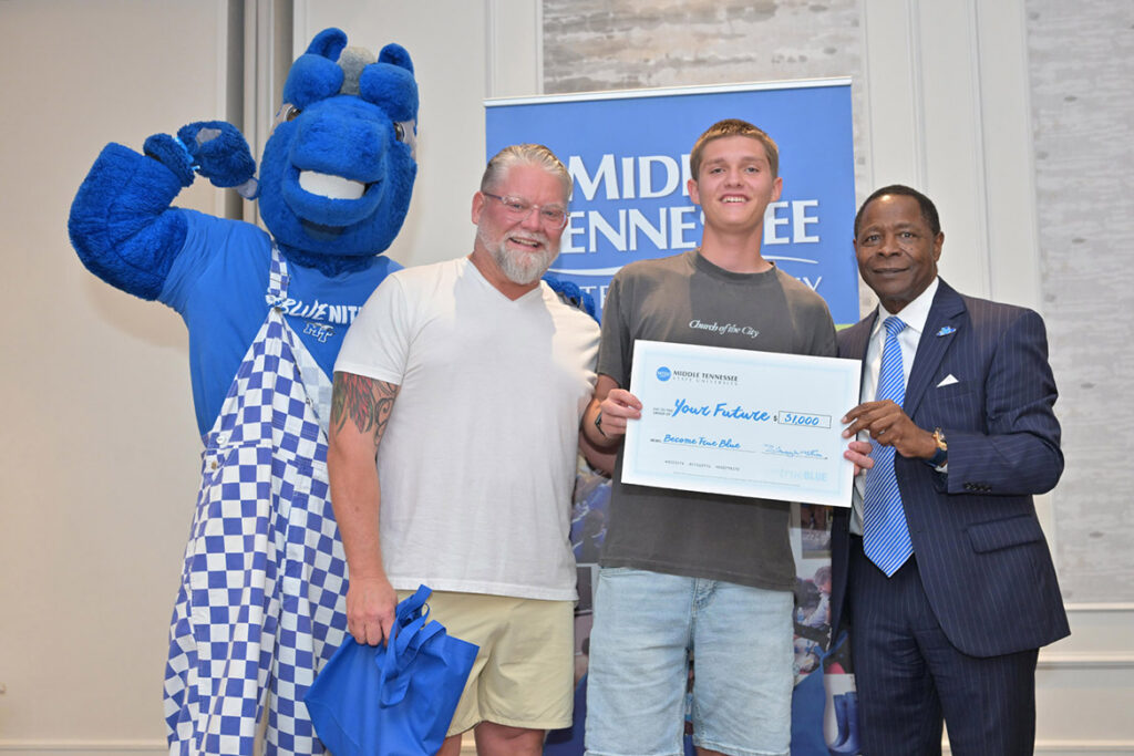 A student, a parent, the university president and the university mascot smile at the camera, holding a ceremonial check.