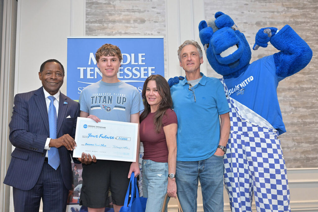 A student, two parents, the university president and the university mascot smile at the camera, holding a ceremonial check.