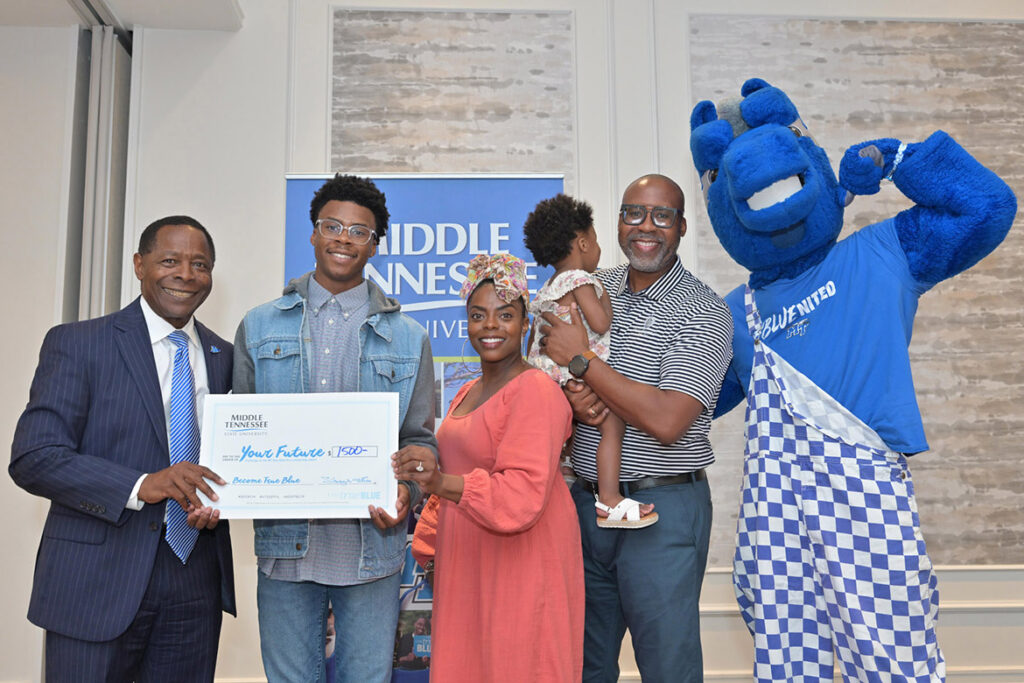 A student, two parents, a baby sibling, the university president and the university mascot smile at the camera, holding a ceremonial check.