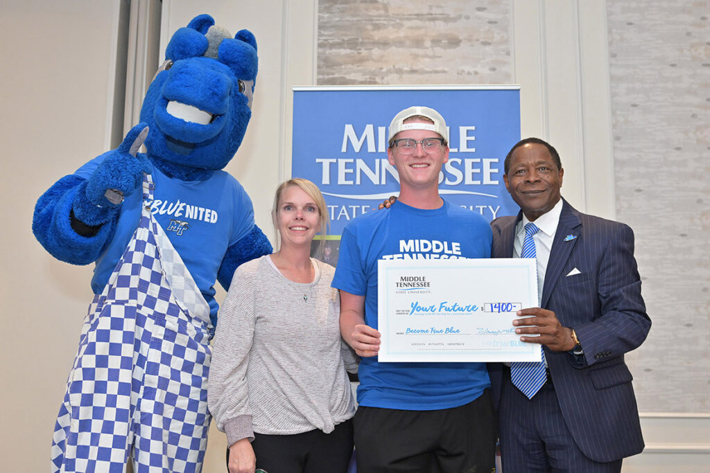 A student, an assistant principal, the university president and the university mascot smile at the camera, holding a ceremonial check.