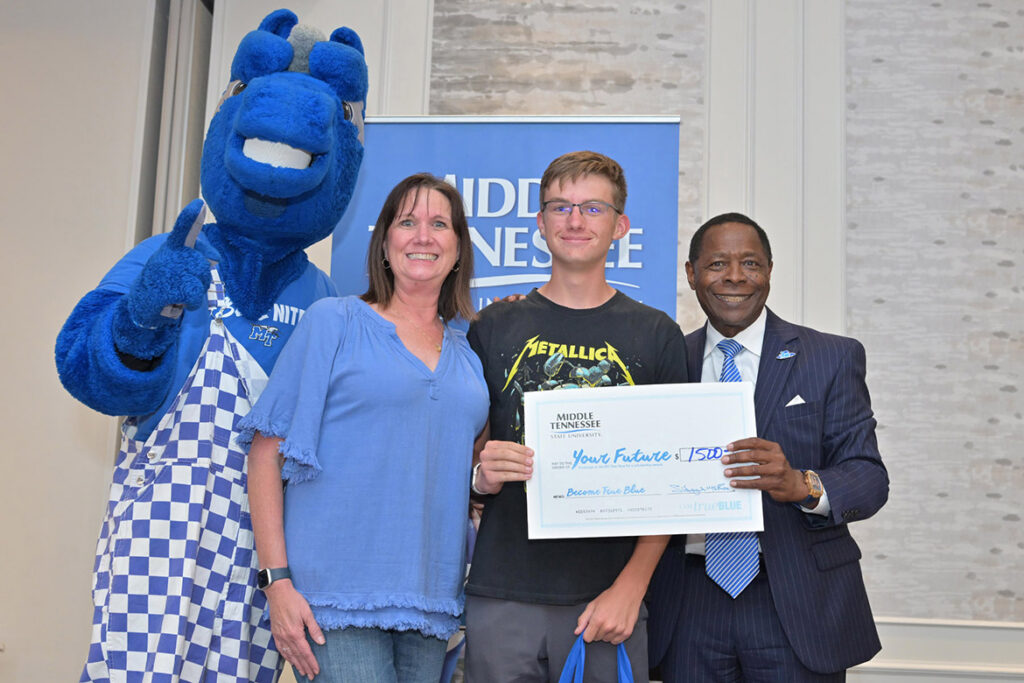 A student, a parent, the university president and the university mascot smile at the camera, holding a ceremonial check.
