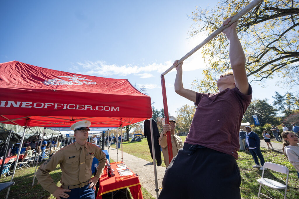 A young civilian wearing a burgundy shirt does pullups while two officers in uniform look on.