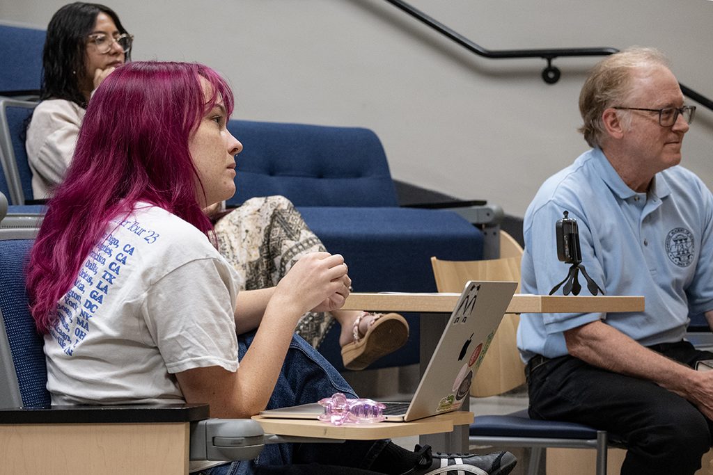 A girl with bright pink hair sits in the audience, listening intently.