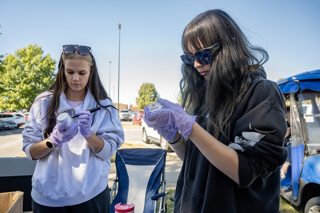 Two female volunteers with long hair, both wearing rubber gloves and sunglasses, use black markers to mark out personal information on prescription bottles.