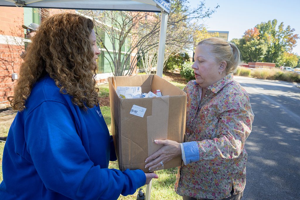A woman with brown, curly hair wearing a blue sweatshirt takes a box being handed to her by a woman with a blond ponytail.